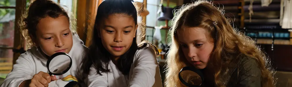 3 人の少女が拡大鏡を使って古い品物を調べています。