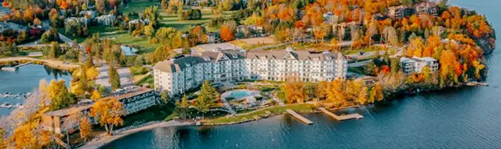 Vista aérea de Deerhurst Resort en Muskoka, enmarcada por las tranquilas aguas del lago y un vibrante bosque otoñal bajo un espectacular cielo rosa-naranja.