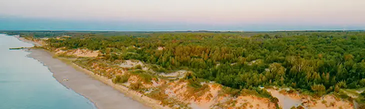 Vista aérea de la playa Ipperwash en Ontario al anochecer, con suaves olas que bañan un largo tramo de costa respaldado por dunas de arena y un denso bosque.