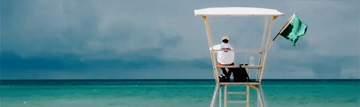 Lifeguard sitting on a tall white chair overlooking the turquoise waters of Grand Bend Beach, with a green flag fluttering in the breeze.
