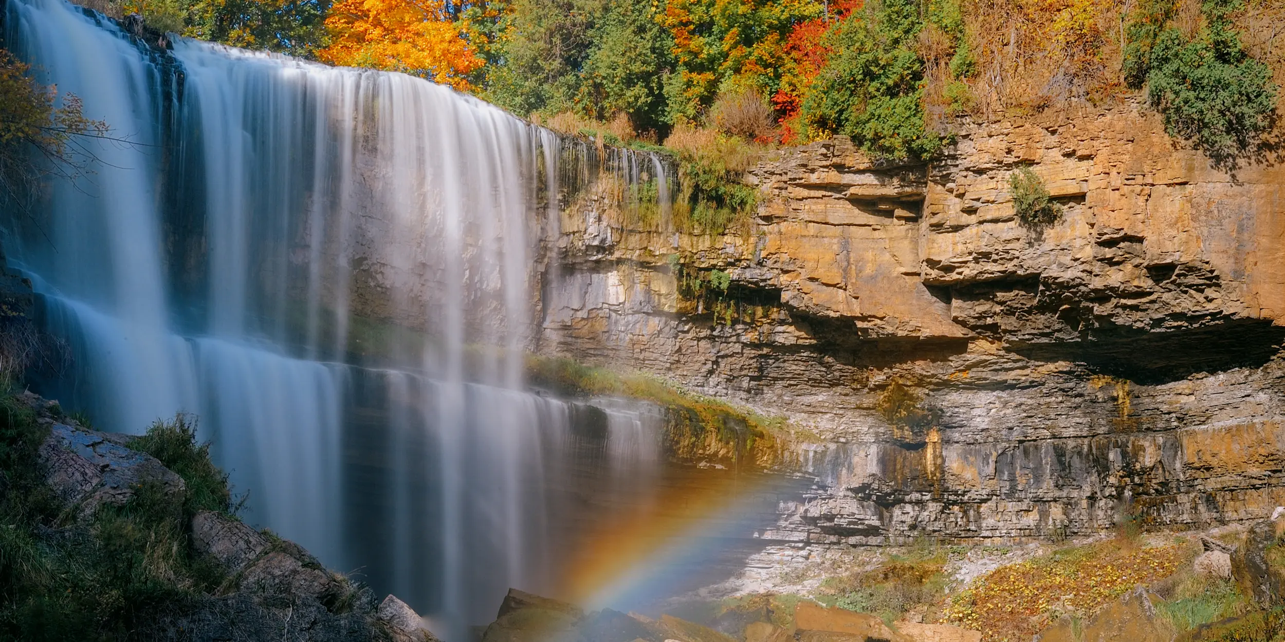  Ein Wasserfall über der felsigen Böschung, umgeben von Herbstlaub und einem Regenbogen im Vordergrund.