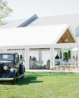 A black vintage car sits on the grass  in front of a large white open air gazebo filled with people sitting on chairs and drinking wine.