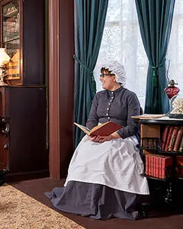 Inside the Bell Homestead, a man and woman dressed in clothes from 1874 smile at each other while sitting in chairs and reading  
