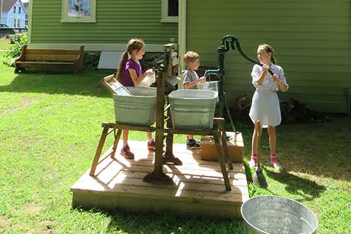 Tres niños llenando cubos de metal con una bomba de agua de estilo antiguo sobre una plataforma de madera.