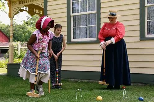 One woman and two young girls dressed in pioneer clothing play croquet next to historic house. 