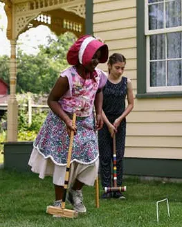 One woman and two young girls dressed in pioneer clothing play croquet next to historic house.