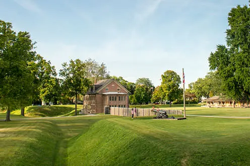 A view of Fort Malden National Historic site from across the grass. It is a a sunny day, people are walking in the distance. 
