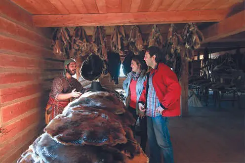 A fur trader displays his wares to a couple of visitors at a trading post