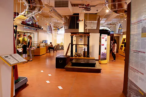 An interior view of the North Bay Museum. There information boards along the walls, a dislpay case to the left and stairs in the middle of the room. There are model planes hanging from the ceiling.