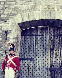 Deux soldats vêtus d'uniformes de la guerre de 1812 se tiennent de chaque côté d'une grande porte en bois menant à un grand fort en pierres.