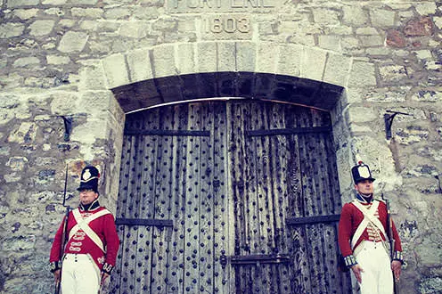 Two male soldiers dressed in War of 1812 uniforms standing on either side of a large wooden door leading into a large stone fort. 