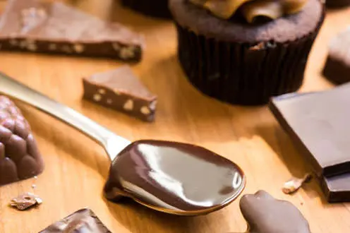 Un primer plano de una cuchara de metal cubierta de chocolate, rodeada por una magdalena de chocolate y una corteza de chocolate.