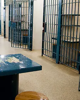 A hallway lined with open jail cells inside the Historic SDG Jail. On the left hand side are blue metal tables with stools.