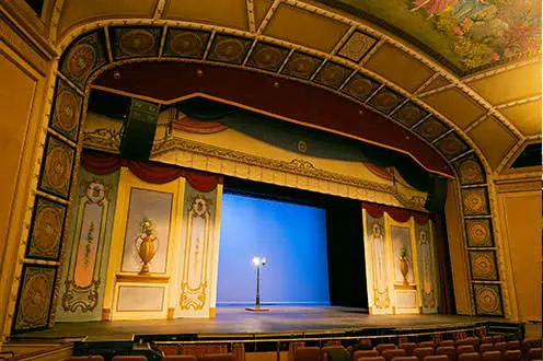 Un teatro interior más pequeño cuyo escenario está rodeado de azulejos y pinturas ornamentados. Una única lámpara de pie se encuentra en el centro del escenario.