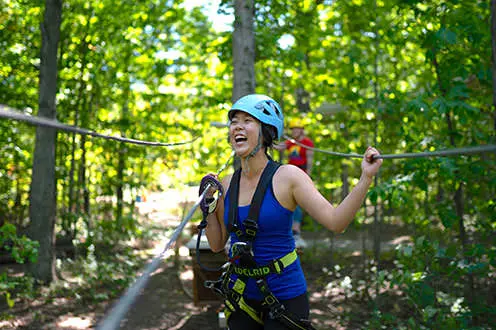 A woman wearing a harness laughs while attached to ziplines in a forest.   