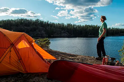晴れた日に湖のほとりにテントとカヌーの横に立っている男性。遠くの海岸線は岩の崖で構成されています。