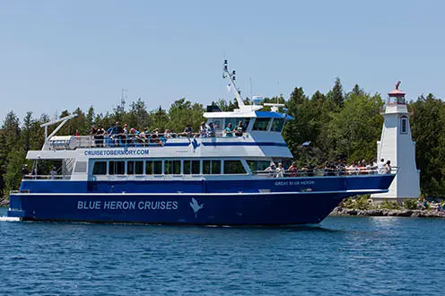 Sur un grand bateau, des gens ayant pris place à l'avant et sur le pont supérieur naviguent près d'un phare.  Sur la coque du bateau, on peut lire "Blue Heron Cruises".