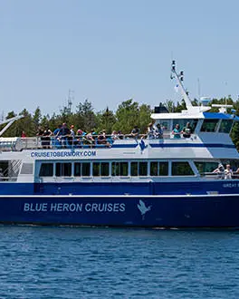 A large boat with people on the front and top deck sailing by a lighthouse. The boat has "Blue Heron Cruises" written on the side.