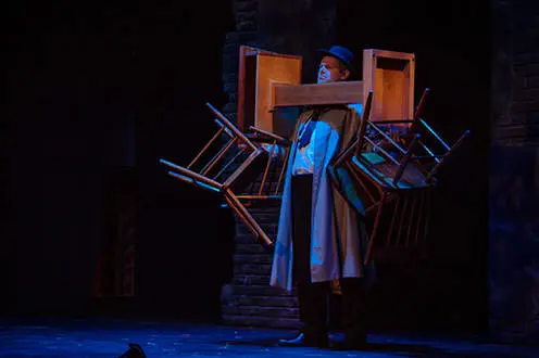 트렌치코트와 모자를 쓴 남자가 파란 스포트라이트를 받고 서 있다. 그는 각 손에 두 개의 의자를 들고 의자와 받침대의 균형을 잡고 있습니다.