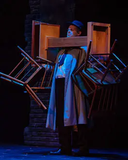 Un hombre vestido con una gabardina y un sombrero está de pie con un foco azul brillando sobre él. Sostiene dos sillas en cada mano y equilibra un pedestal con las sillas.
