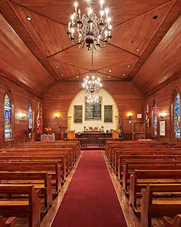 The inside of a chapel covered with wooden panelling and three stained glass windows on each side. An aisle separates two rows of wooden pews and three chandeliers are hanging above. 