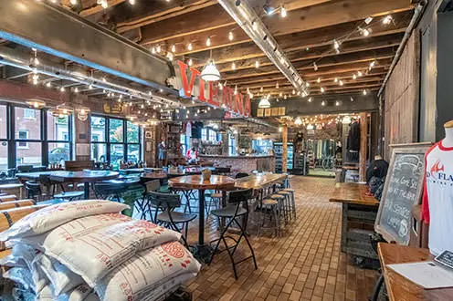 A large room with open celings and string lights. Many different sizes of tables fill the room along with bags of barley and mannequins wearing t-shirts. The word VIENNA is displayed horizontally along a wooden beam close to the ceiling 