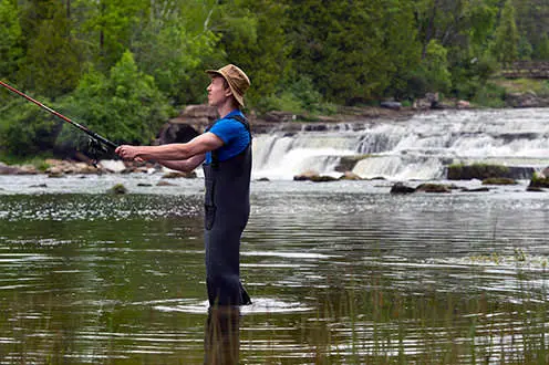 湖のほとりにウェーダーをかぶった男が釣り竿を投げている。遠くに滝があります。