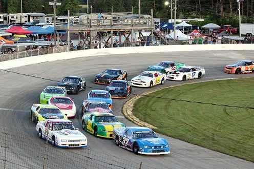 楕円形の競馬場の曲がり角を高速で走る15台のレースカー。背景にはテントがあり、木製の展望台には観客が立っています。