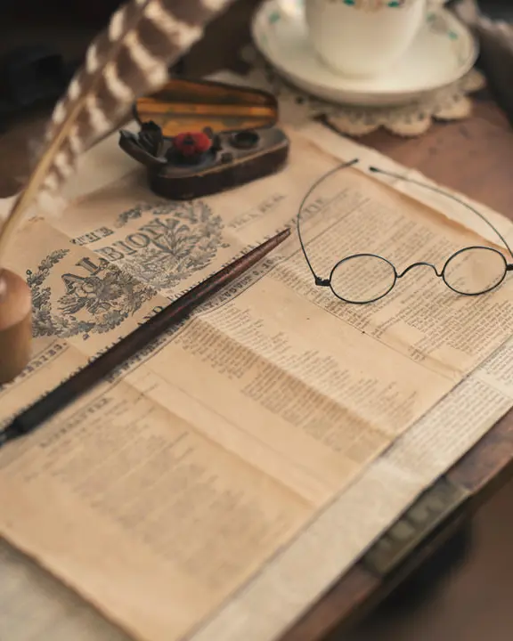 Table avec tasse et soucoupe de porcelaine à côté de la première page d'un journal historique avec des stylos et des lunettes de lecture d'époque sur le dessus.
