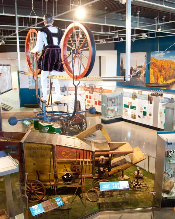 Rücken eines Mannes, der hoch zwischen 2 großen historischen Wagenrädern steht und von Pioniermöbeln und Fotos an den Wänden umgeben ist