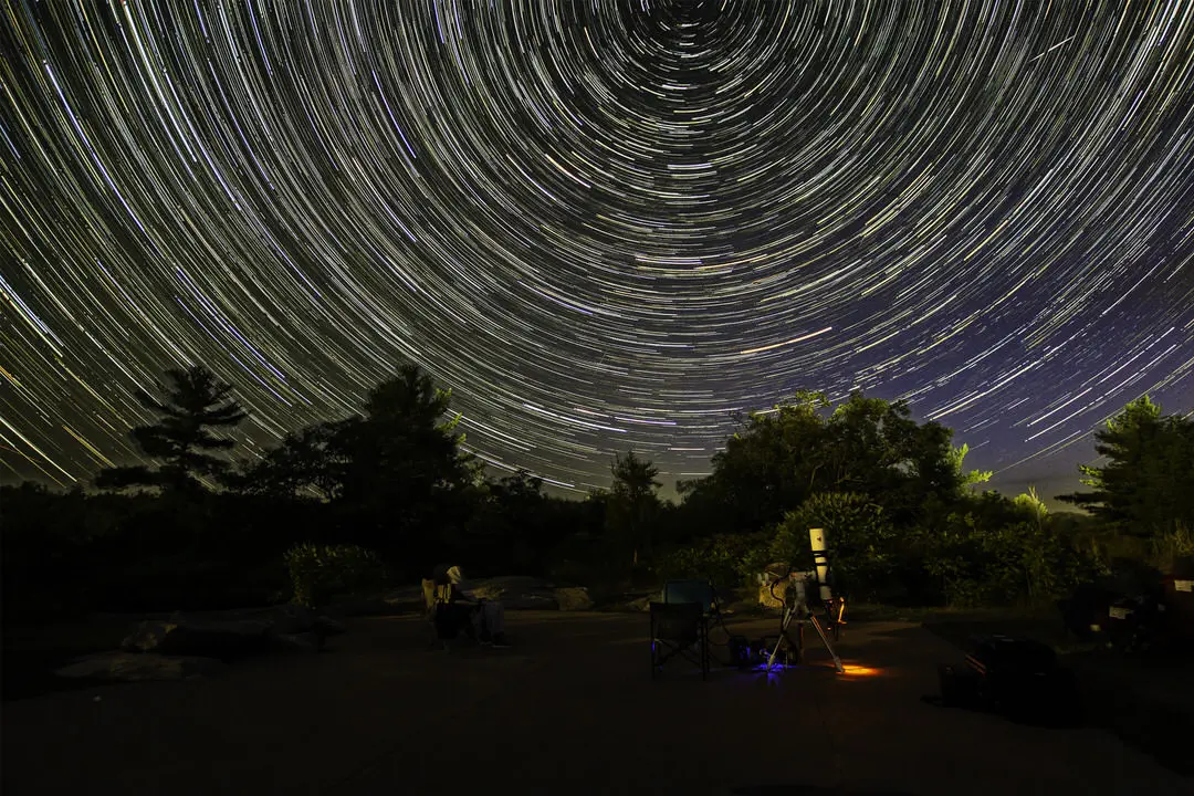 何百もの輝く平行な円形の星の小道がある暗い空に圧倒される背景の森に面した公共の望遠鏡