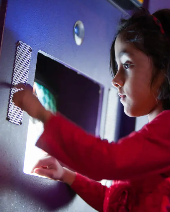 Small child standing in front of recessed computer screen pressing dots on small metal plate on the wall to her left