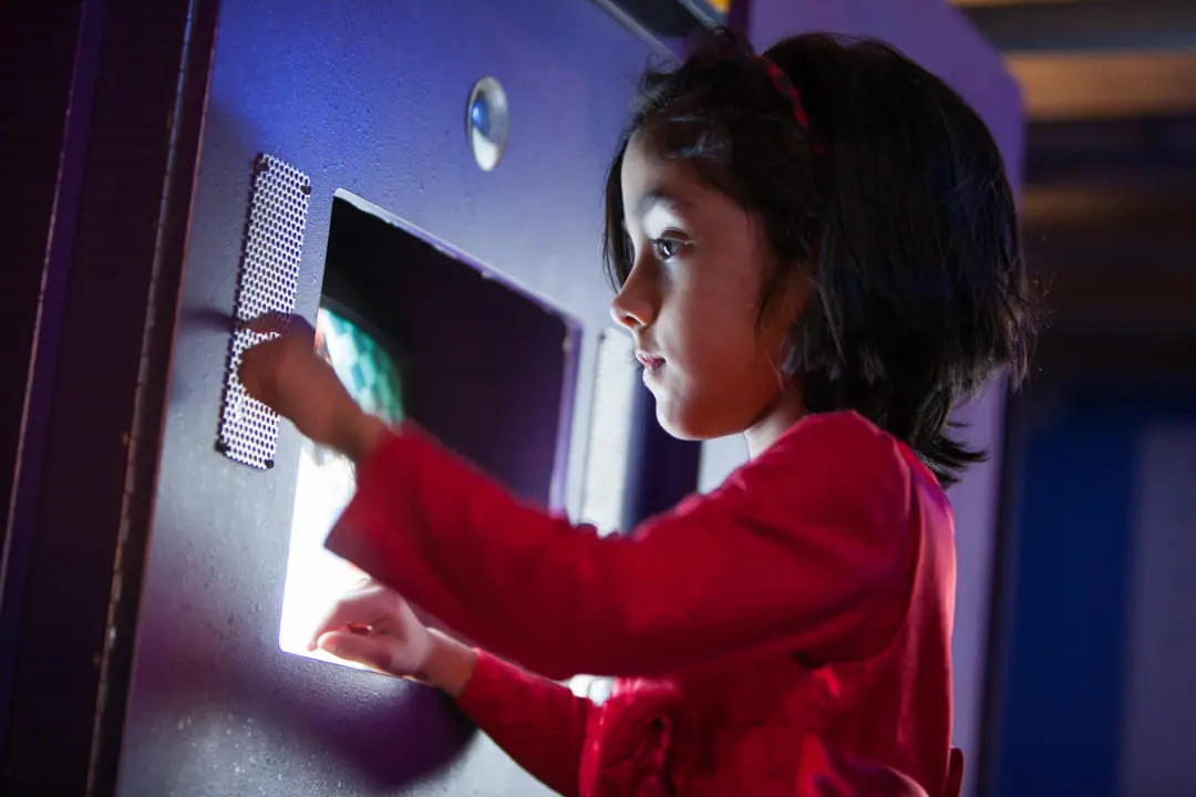 Small child standing in front of recessed computer screen pressing dots on small metal plate on the wall to her left