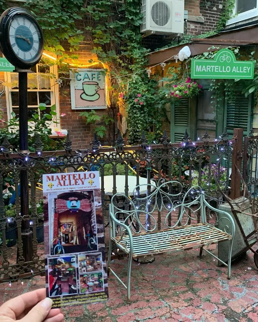Affiche de Martello Alley à côté d'un banc vert rouillé et d'une brouette sur une ancienne promenade de brique avec un café extérieur derrière la clôture.