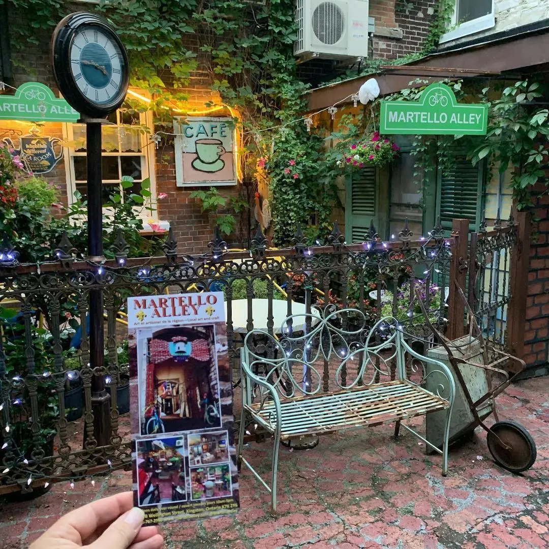Affiche de Martello Alley à côté d'un banc vert rouillé et d'une brouette sur une ancienne promenade de brique avec un café extérieur derrière la clôture.