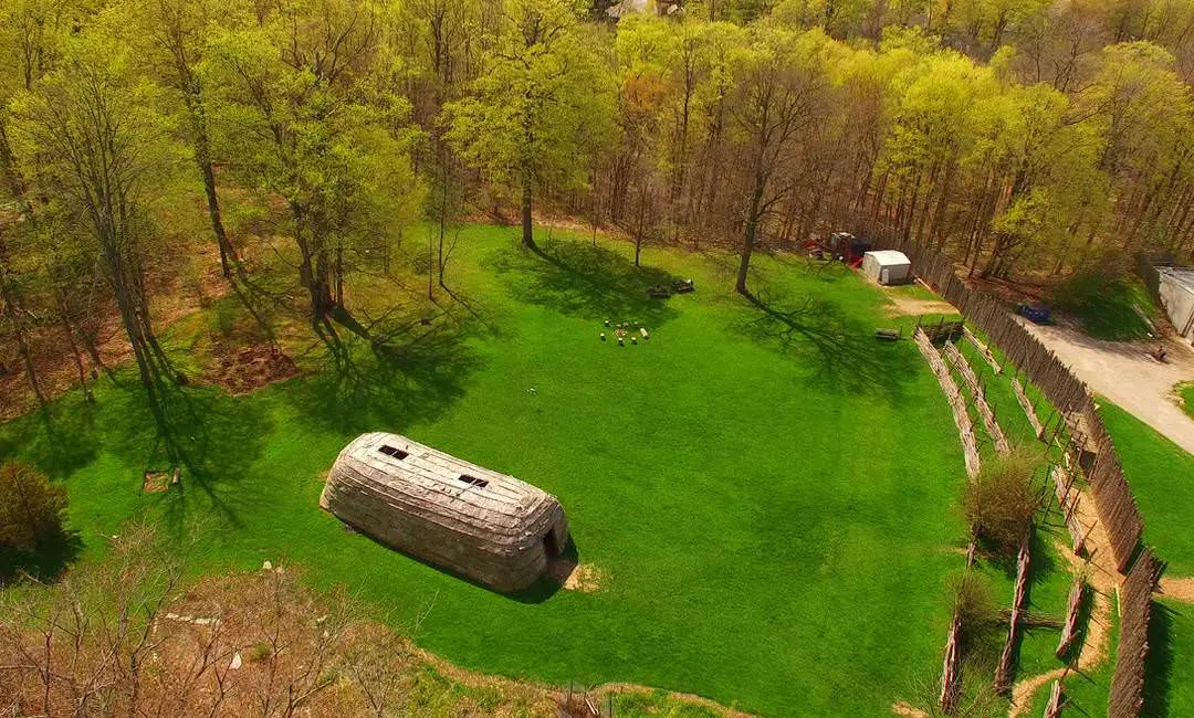 Aboriginal brown longhouse in middle of large green lawn surrounded by forest with wood palisades along one side
