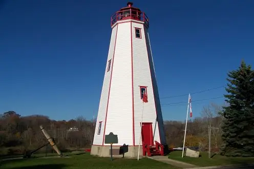 Green lawn with very tall white lighthouse with red doorway and front steps and balcony and with forest beyond