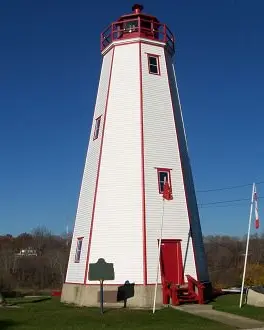 Pelouse verte avec un très grand phare blanc et porte d'entrée rouge ainsi que des marches avant et un balcon avec une forêt plus loin.