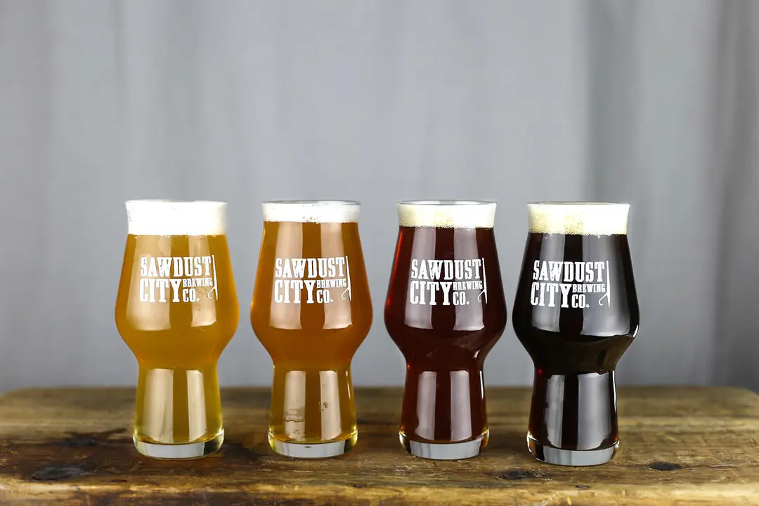 4 glasses each containing an ever darker colour of beer lined up on old wood table and each printed with company name