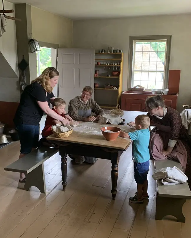 長い開拓者のドレスを着た3人の女性と2人が台所のテーブルで、現代の女性と2人の小さな男の子がパン生地をこねるのを手伝っています