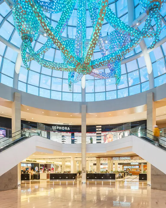 Centre commercial circulaire à deux étages avec vitrines de magasins et éclairé par de nombreuses lumières miniatures colorées sur des tubes suspendus depuis un immense plafond en dôme de verre.