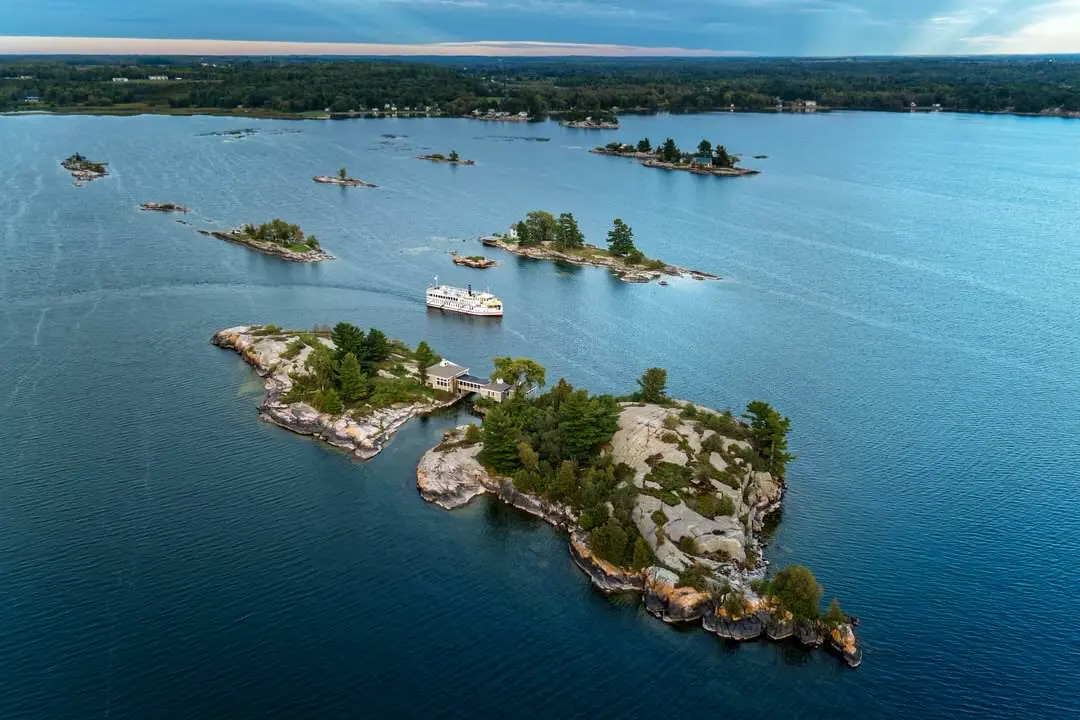 Large rivière avec deux îles, une grosse et une petite, où se trouvent des arbres et des bâtiments ainsi qu'un traversier qui effectue la traversée depuis la rive opposée au loin.