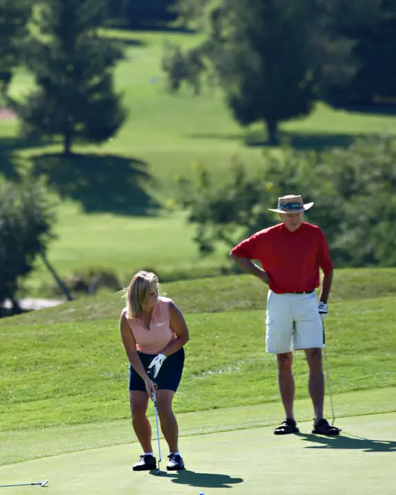 Woman on golf course lining up her golf club for a shot with man standing watching with backdrop of green grass and evergreens