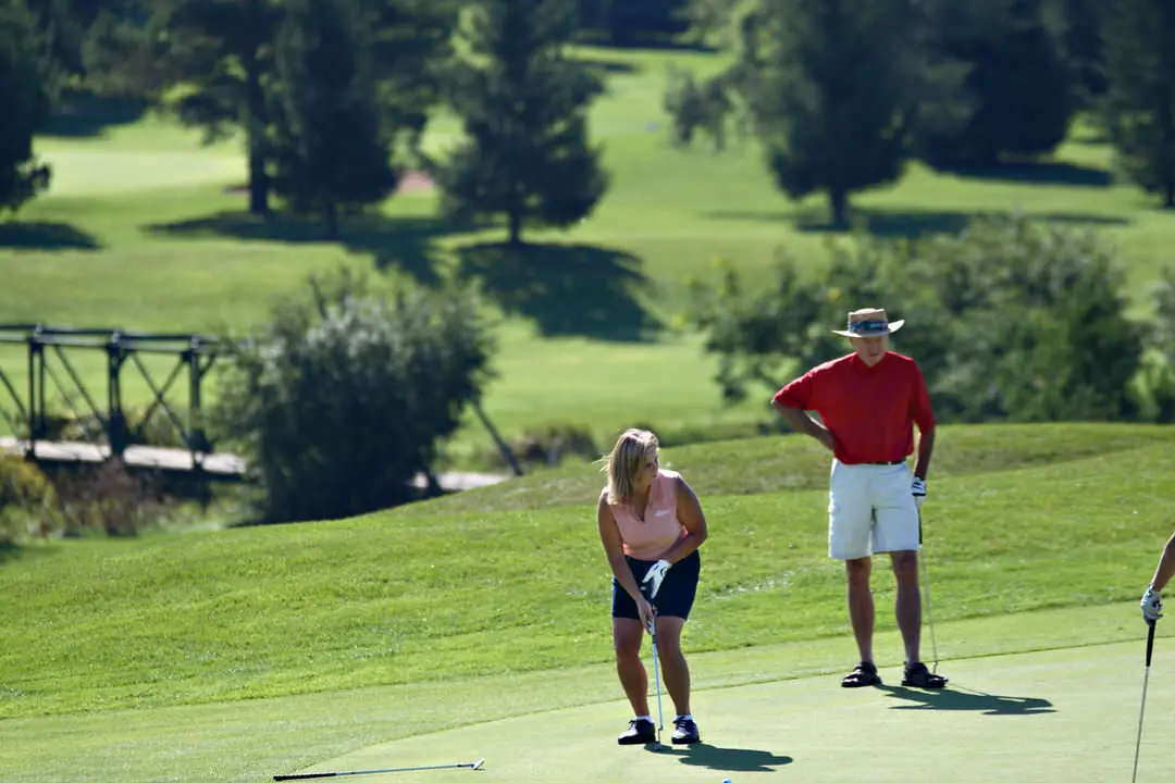 Une femme sur un terrain de golf alignant son bâton pour frapper la balle alors qu'un homme est debout et regarde avec en arrière-plan de l'herbe verte et des conifères.
