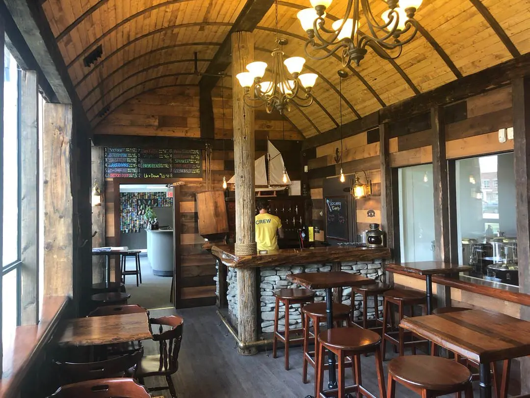 Rustic wood restaurant with wood tables and stools and chairs and bar at back corner with employee facing shelves of bottles
