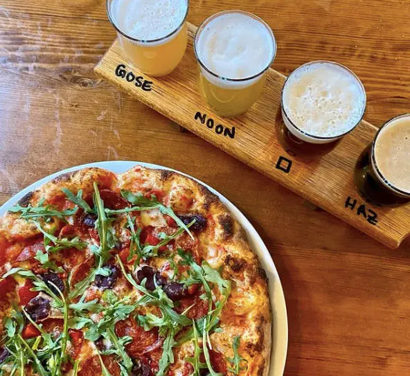 Pizza entière sur une assiette à côté d'un plateau sur lequel se trouve une rangée de quatre verres de bière de différentes couleurs avec le type de bière imprimé sur le plateau.