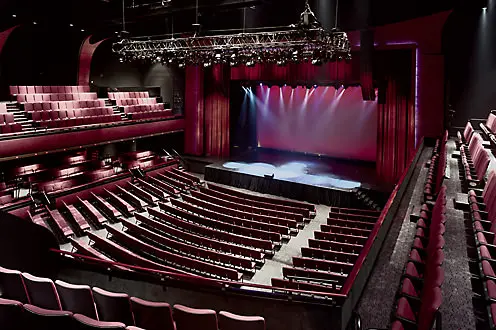 Filas de asientos de color vino en el teatro tanto en el piso principal como en los balcones que dan al escenario vacío con toda la sala tenuemente iluminada
