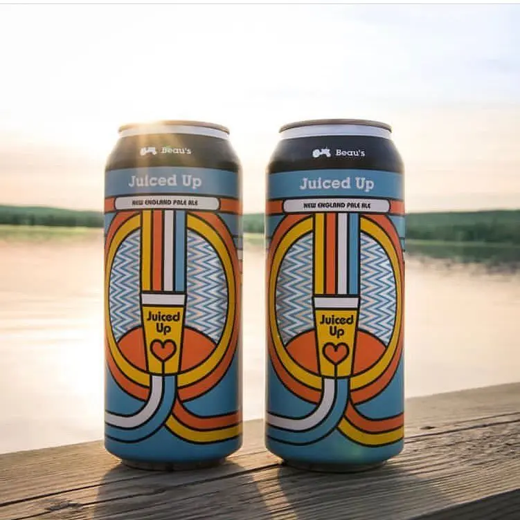 Close up of two colourful Beau's Juiced Up beer cans sitting on a table with the lake in the background