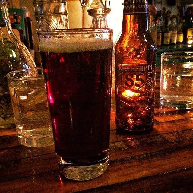 Close up of a glass of a dark ale beside a bottle of beer sitting a top a bar