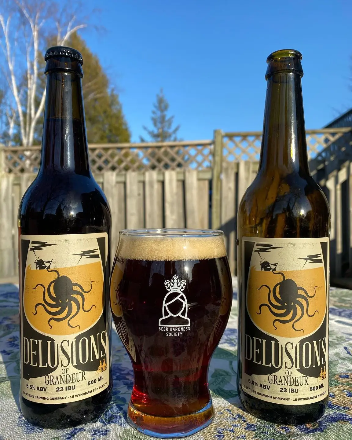 A glass of dark ale sitting between two beer bottles in a backyard setting